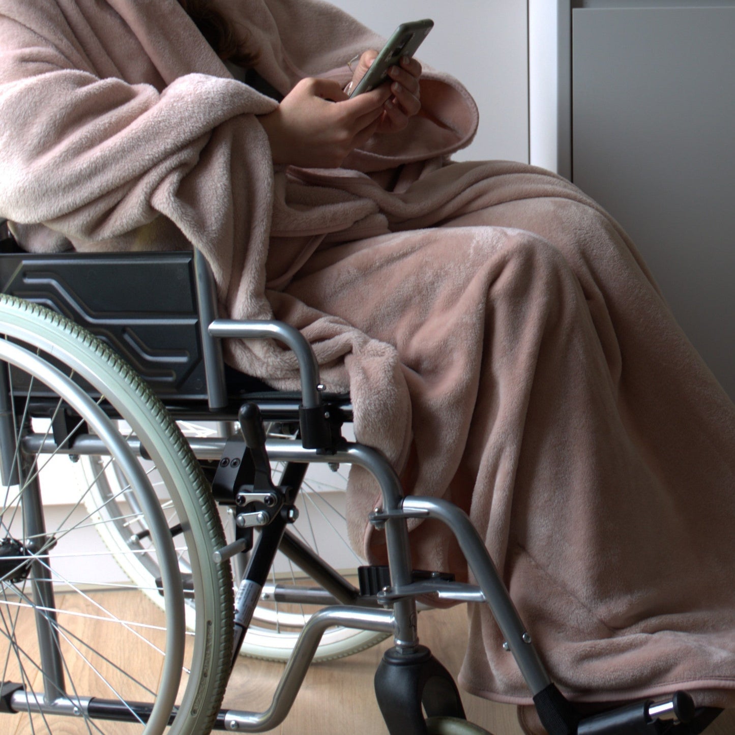 Person sitting in a wheelchair indoors, wearing a Snhug Sit sitting blanket and using a smartphone, demonstrating accessible comfort and warmth.
