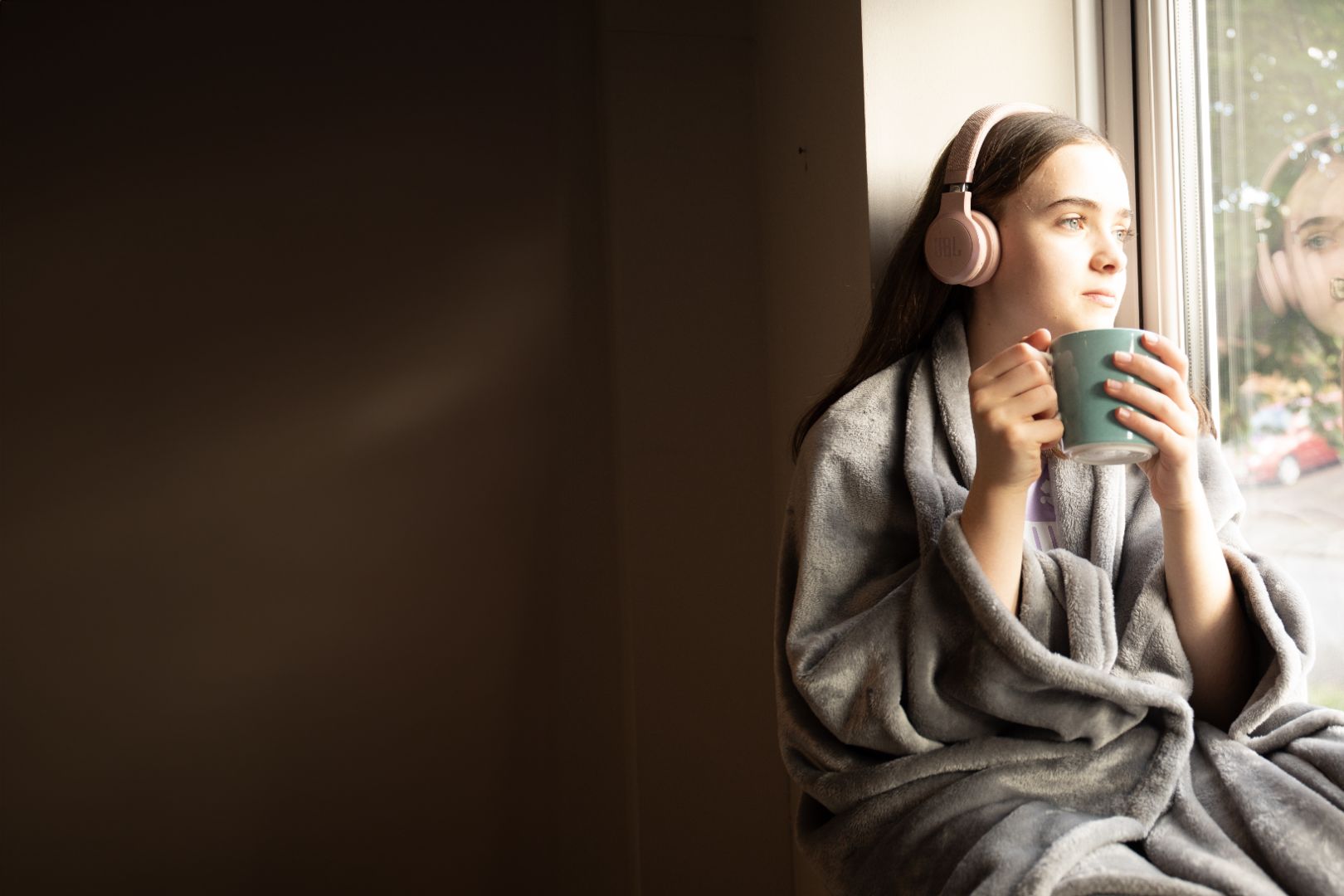 Teen girl wearing a Snhug Sit sitting blanket, listening to music with headphones and holding a mug while relaxing by the window.