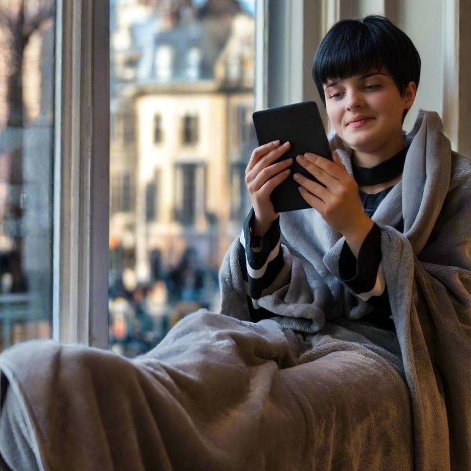 Woman relaxing by the window wearing a Snhug Sit sitting blanket and reading on a tablet, enjoying warmth and comfort indoors.