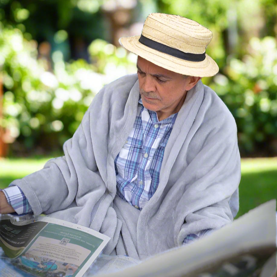 Man sitting outdoors reading a newspaper while wearing a grey Snhug Sit sitting blanket, relaxing in cosy comfort on a cool day.