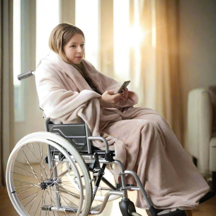Young woman in a wheelchair wrapped in a blush Snhug Sit® blanket, staying warm and comfortable indoors while using her phone—designed for ease, dignity, and support during recovery or reduced mobility.