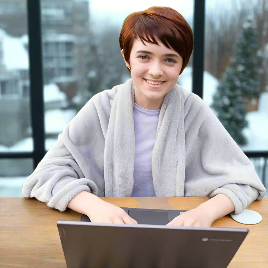 woman wearing a cozy grey Snhug Sit® blanket while using a laptop indoors with snowy trees in the background, enjoying warmth and comfort during winter