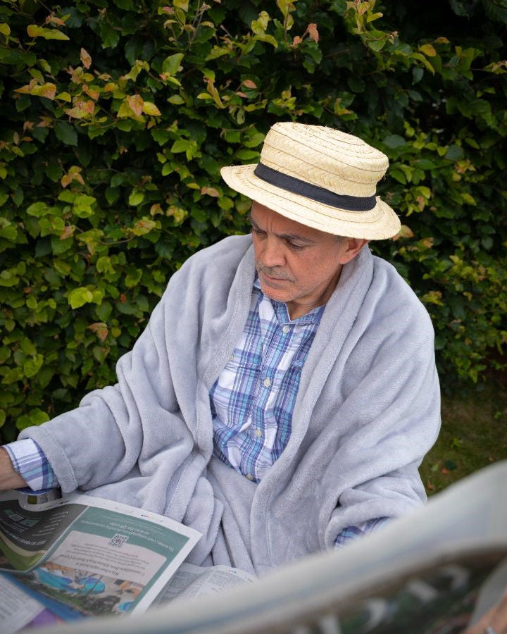 Older man sitting outdoors in a soft grey Snhug Sit® blanket, reading a newspaper in the garden while wearing a straw hat—enjoying warmth, calm, and independence in a relaxing moment